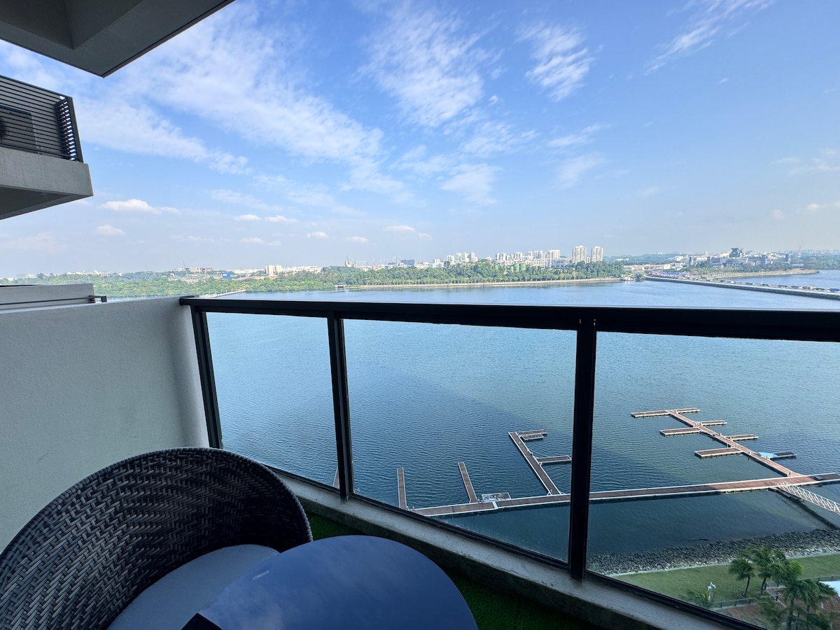 A balcony view is captured, showcasing a serene sea and a distant skyline. The clear blue sky reflects on the water's surface, while a few floating docks are visible in the foreground. A small round table is paired with two woven chairs, placed against the railings.