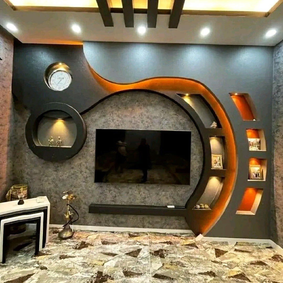 A contemporary living room is showcased with an artistic feature wall that includes a large flat-screen television and decorative shelves. A modern clock is mounted above, while warm lighting highlights the curve of the design. The floor is covered with a textured rug, enhancing the space.