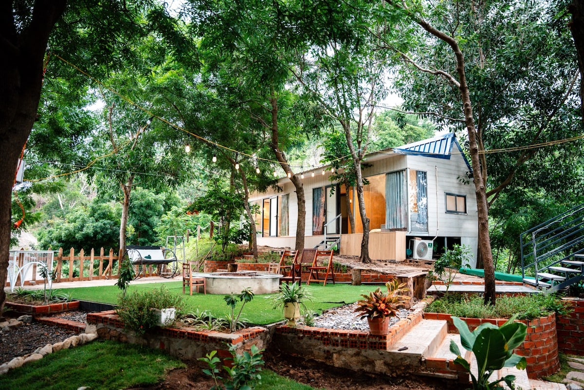 A charming outdoor area features lush greenery and well-maintained landscaping. A circular fire pit is surrounded by wooden chairs, with a patio area visible. The dwelling, characterized by large windows and a blue roof, is nestled among trees, creating a serene environment.