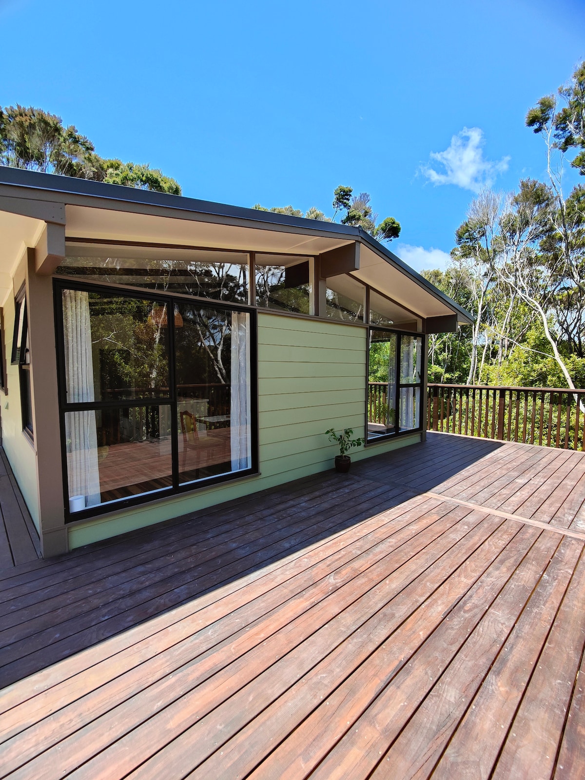 A modern home exterior is highlighted by large glass windows that invite natural light. A wooden deck extends from the house, offering ample space for outdoor relaxation. Lush greenery surrounds the area, creating a natural backdrop against a clear blue sky.