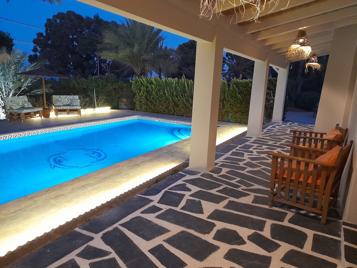 An outdoor area features a tranquil swimming pool illuminated in soft blue light, surrounded by palm trees. A patterned stone pathway leads to comfortable seating arrangements, with cushioned chairs positioned under a wooden overhang. Natural landscaping provides privacy from neighboring areas.