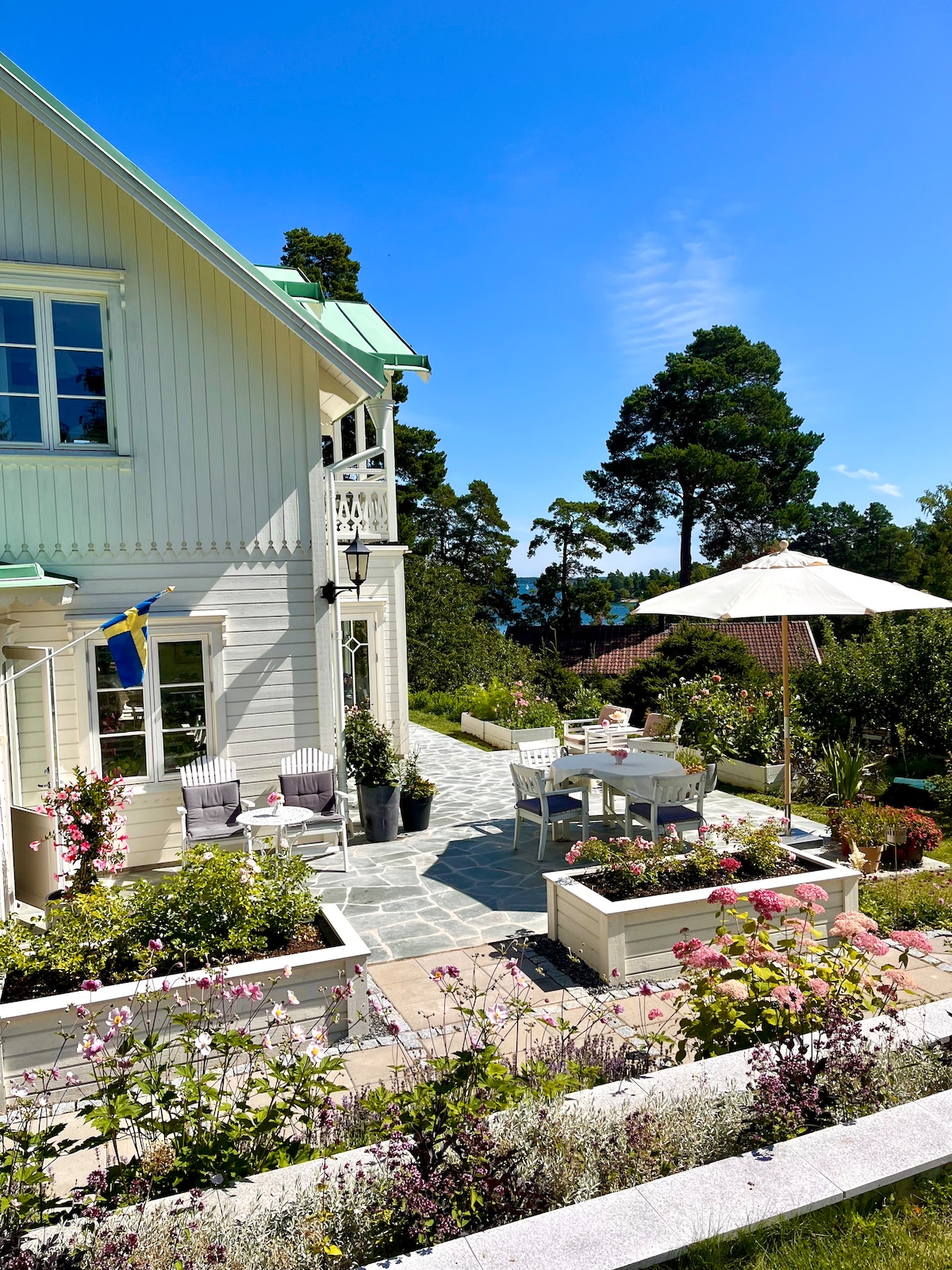 A serene outdoor patio area is surrounded by vibrant flowers. A white dining table with matching chairs is sheltered under an umbrella. The building’s light-colored wood contrasts with the greenery of trees in the background, providing a tranquil space for relaxation.