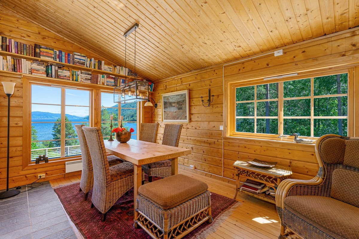 A bright dining area features a wooden table surrounded by woven chairs. A large window offers a view of the landscapes outside, while bookshelves filled with books line the wall. Natural light fills the space, enhancing the wooden interior elements.