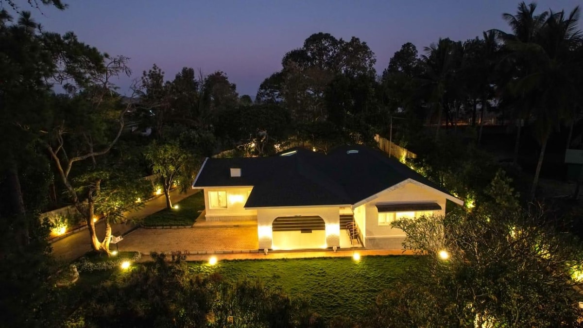 A well-lit modern house is surrounded by lush greenery, creating a tranquil environment. Outdoor lighting highlights the landscaped area, while the home features large windows that invite natural light. The evening sky casts a serene backdrop over the property.