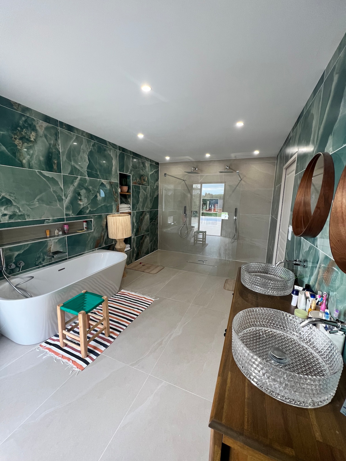A spacious bathroom features elegant green marble wall tiles and an open shower area. A freestanding bathtub is centered on the floor, while double sinks with woven bowls are positioned on a wooden vanity. A patterned rug adds a touch of warmth to the space.