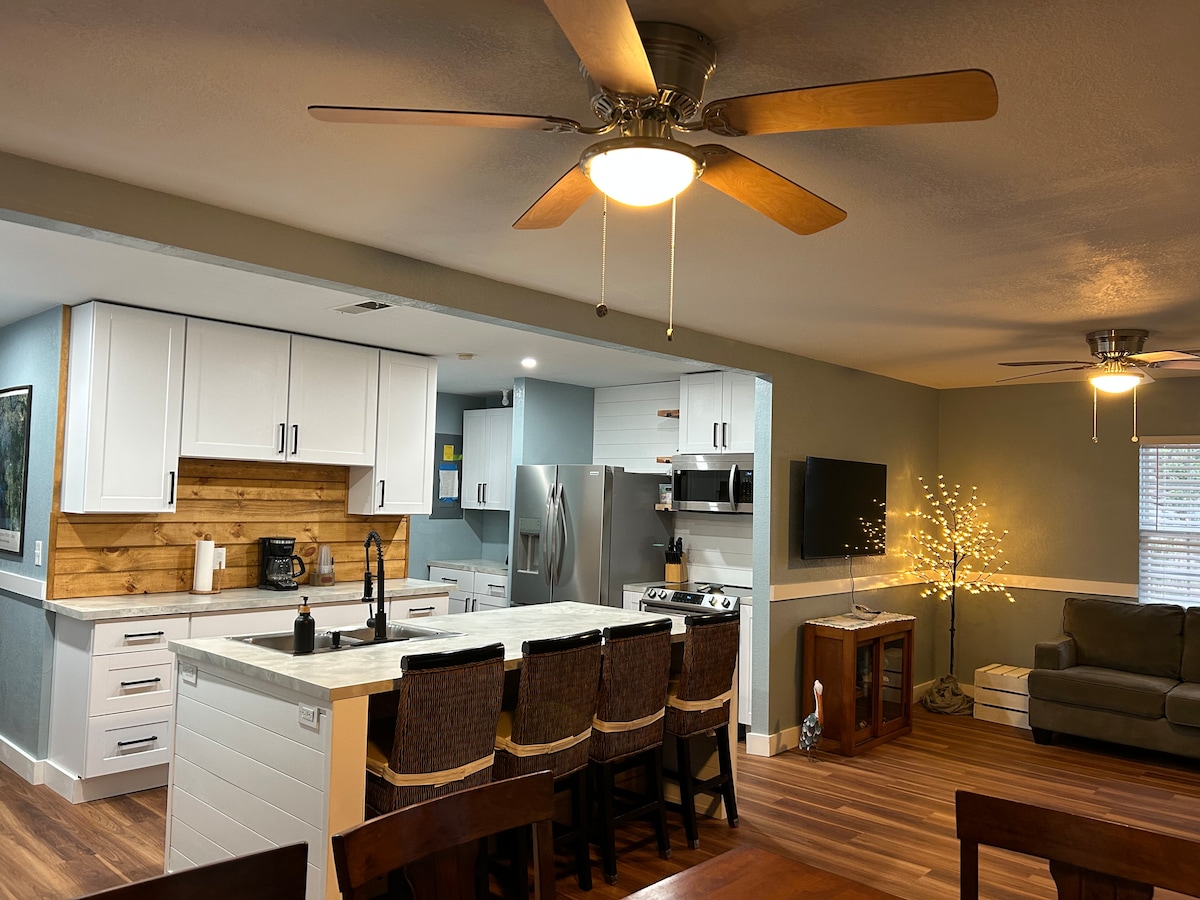 A spacious kitchen area is showcased, featuring modern white cabinetry and a wooden accent wall. A large island with seating for several guests is visible, alongside stainless steel appliances. The adjoining living area includes a cozy couch, a television, and a softly glowing lighted decor element.