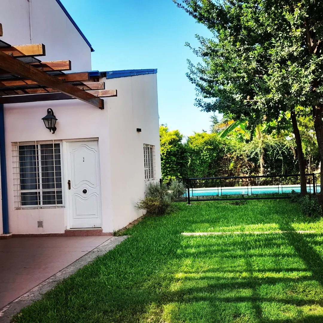 A well-maintained lawn extends beside a white house featuring a prominent front door and secure windows. The patio area is sheltered by wooden beams, while a pool is visible in the background, offering a glimpse of the natural greenery surrounding the property.
