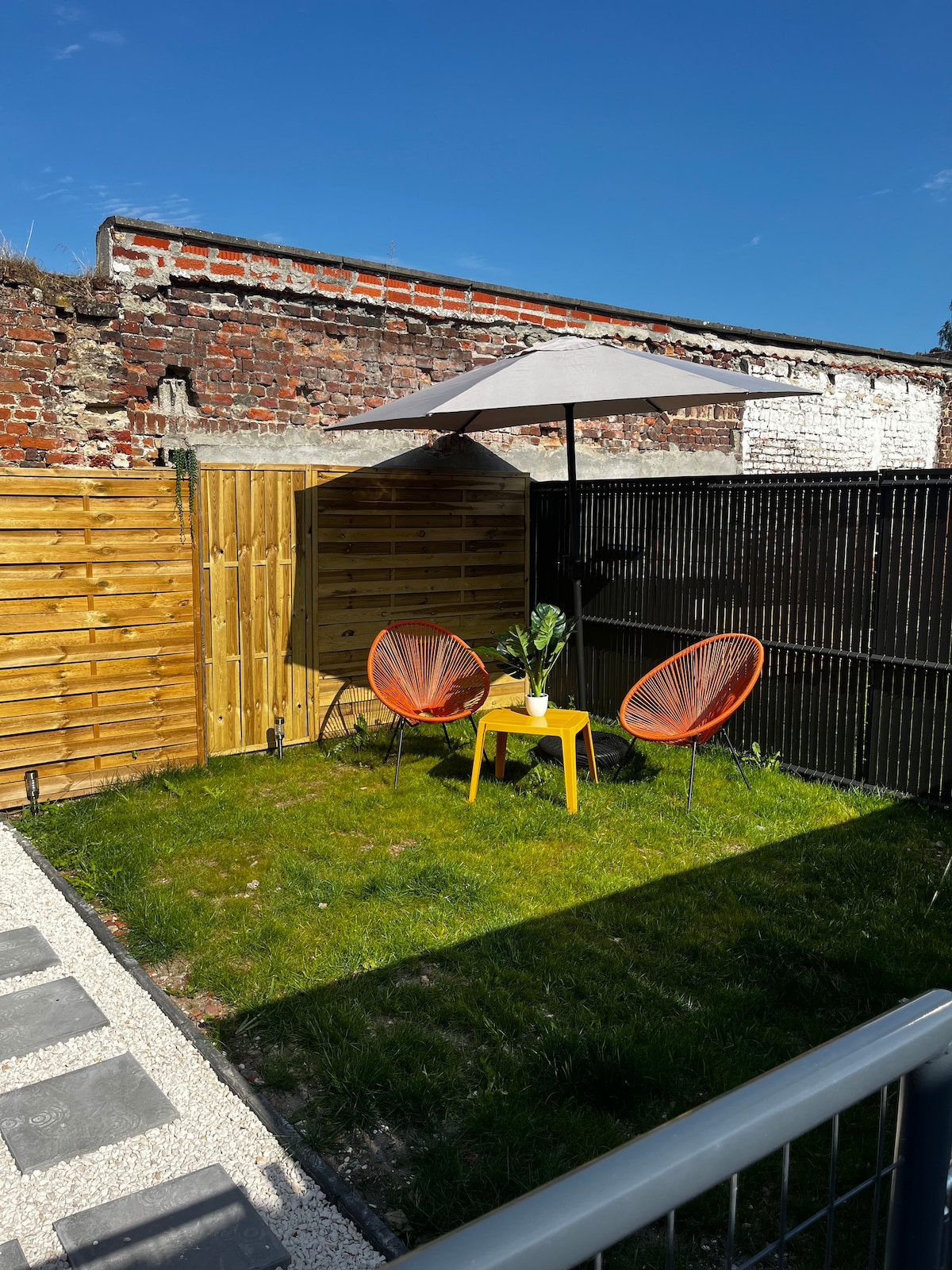 An outdoor space is presented, featuring a green lawn bordered by wooden fencing. Two orange chairs are placed around a small yellow table. A beige umbrella provides shade, and a pathway of stones leads to the area, against a backdrop of rustic brick walls.