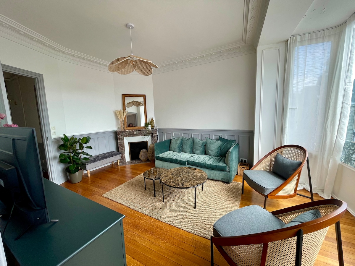 A spacious living area features a teal sofa and two woven chairs positioned around a circular coffee table. A flat-screen television is situated on a low console. Natural light filters in through sheer curtains, complementing the wooden flooring and decorative fireplace.