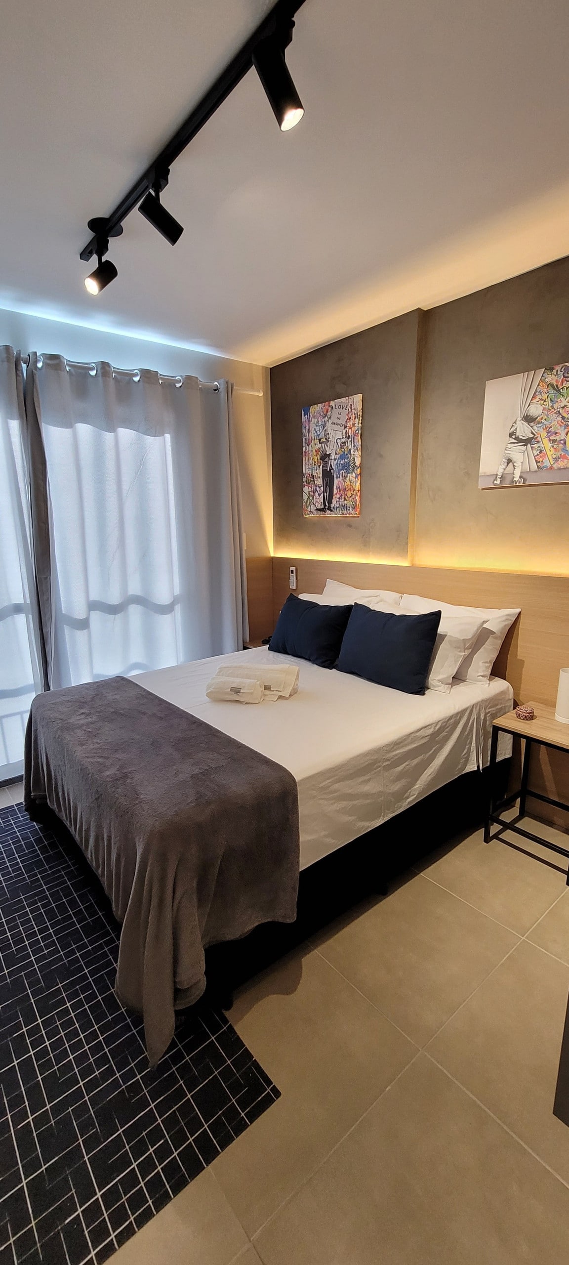 A clean and modern sleeping area features a spacious bed with dark blue and gray accents. Two pieces of colorful wall art are displayed above the bed, with soft lighting illuminating the room. Natural light enters through sheer curtains, creating a serene atmosphere.