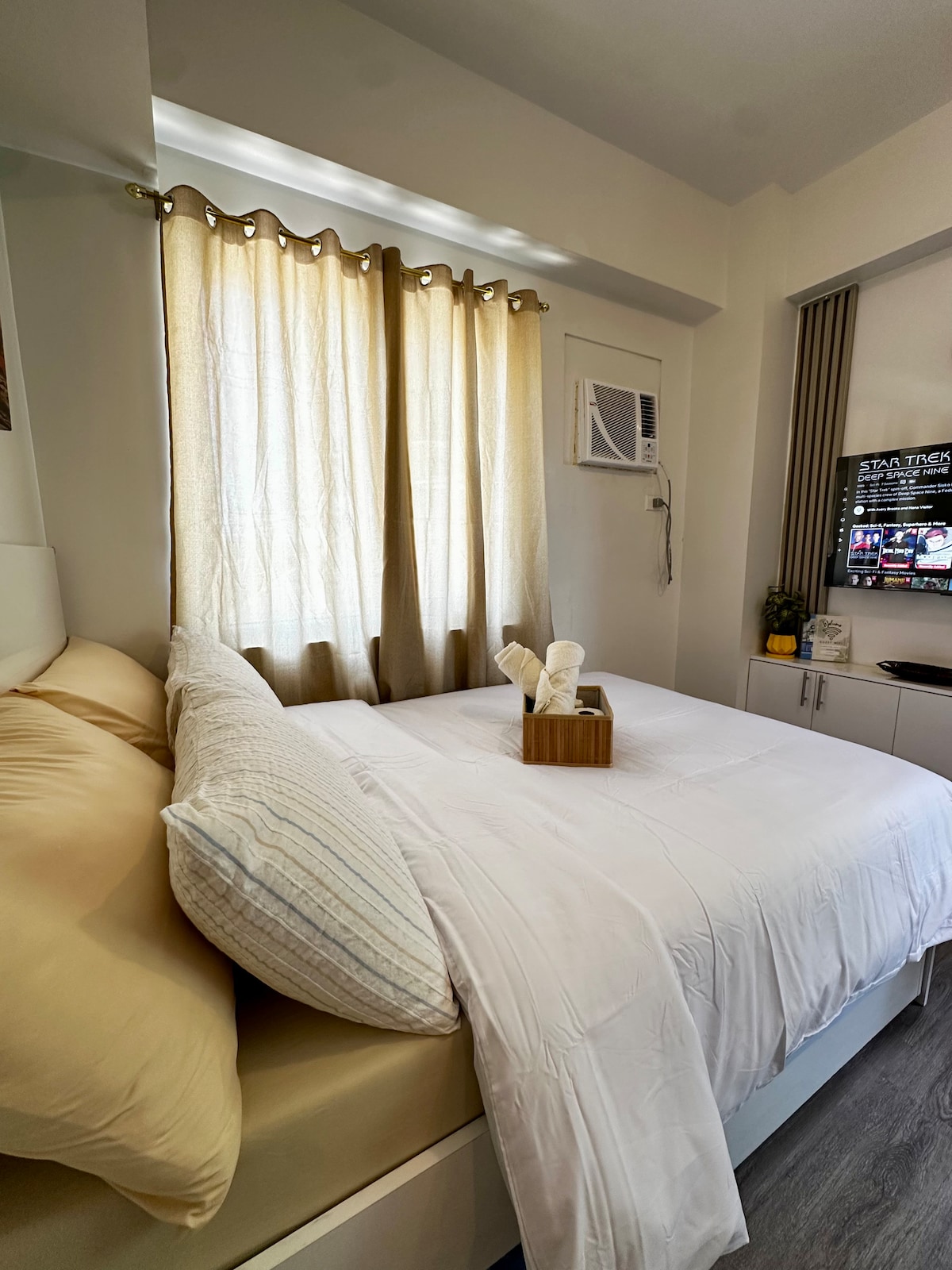 A cozy sleeping area is featured with a spacious bed layered in neutral-toned bedding. The light curtains allow soft natural light to filter in. A mounted TV is visible across from the bed, and a wooden tray with rolled towels is placed at the foot.