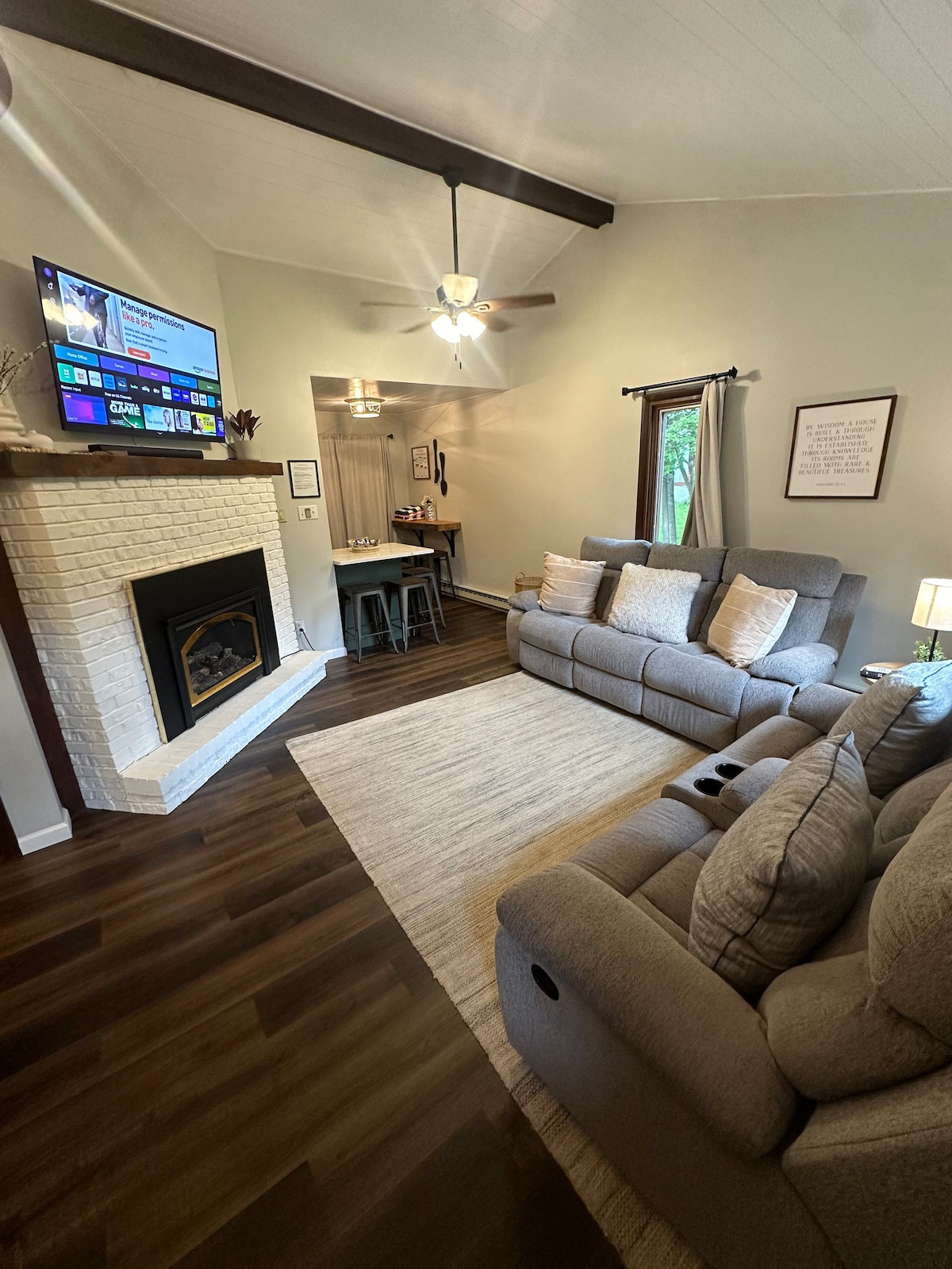 A comfortable living space features a light-colored area rug covering dark flooring. A sectional sofa, equipped with built-in cup holders, faces a mounted TV above a white brick fireplace. Natural light enters through nearby windows, creating a welcoming environment.