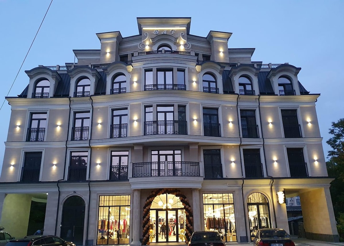 A modern five-story building showcases elegant architectural details with arched windows and balconies. Soft lighting highlights the façade, enhancing its contemporary design. The ground floor features large glass storefronts, providing a view of the interior retail space.