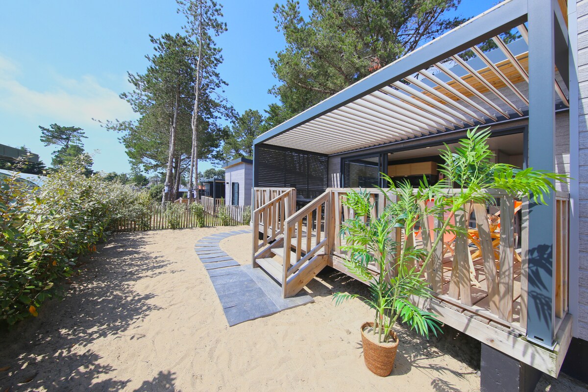 The exterior of the cottage is featured, showcasing a welcoming terrace with a pergola. A clear pathway leads to the entrance, surrounded by garden areas with sandy ground and green foliage. Sunlight filters through the trees, highlighting the outdoor space.