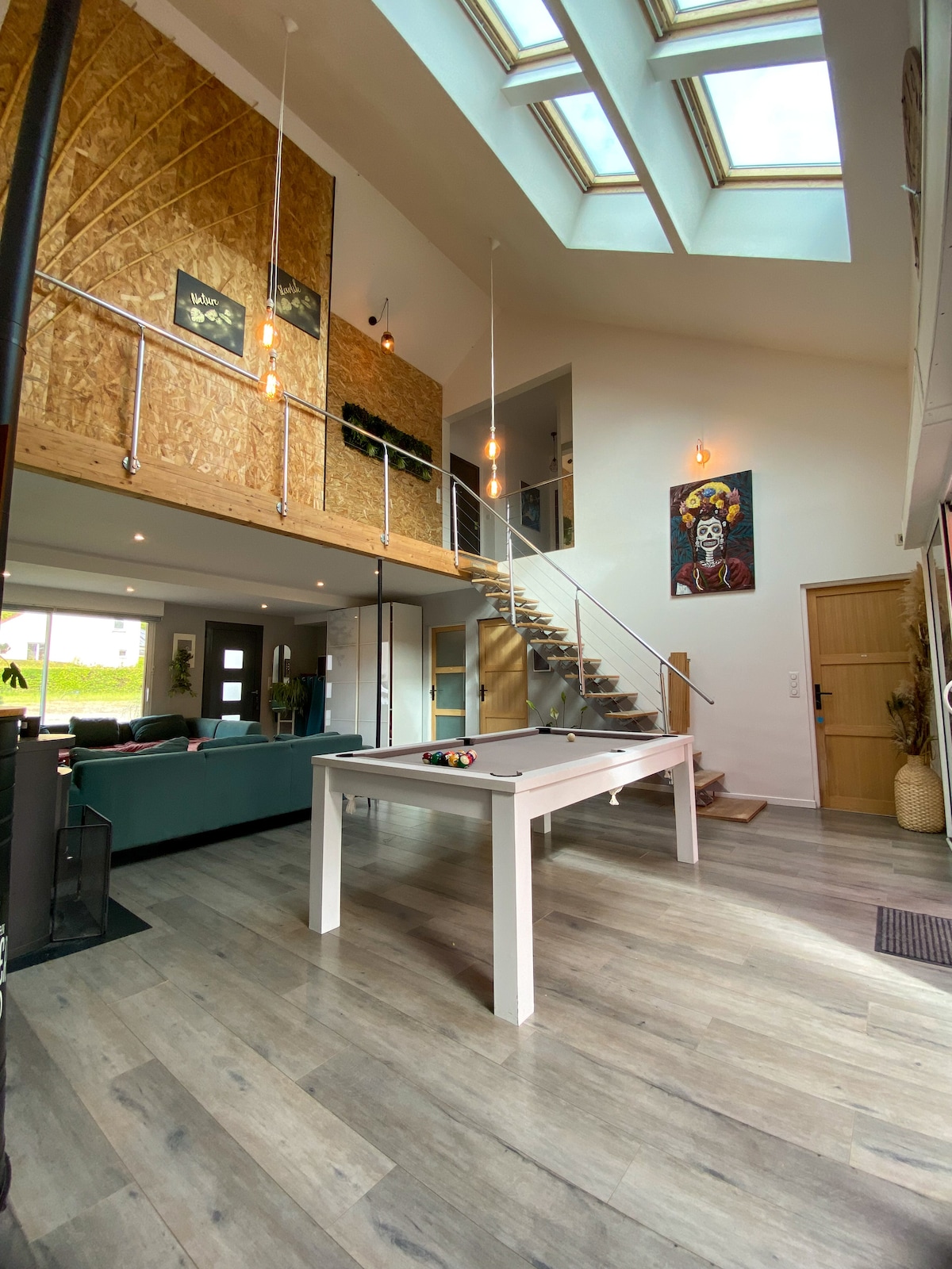 A spacious living area is highlighted by tall ceilings and large skylights allowing natural light to fill the space. A sleek billiards table sits in the center, surrounded by contemporary décor. A staircase leads to the upper level, with a glass railing enhancing the openness.