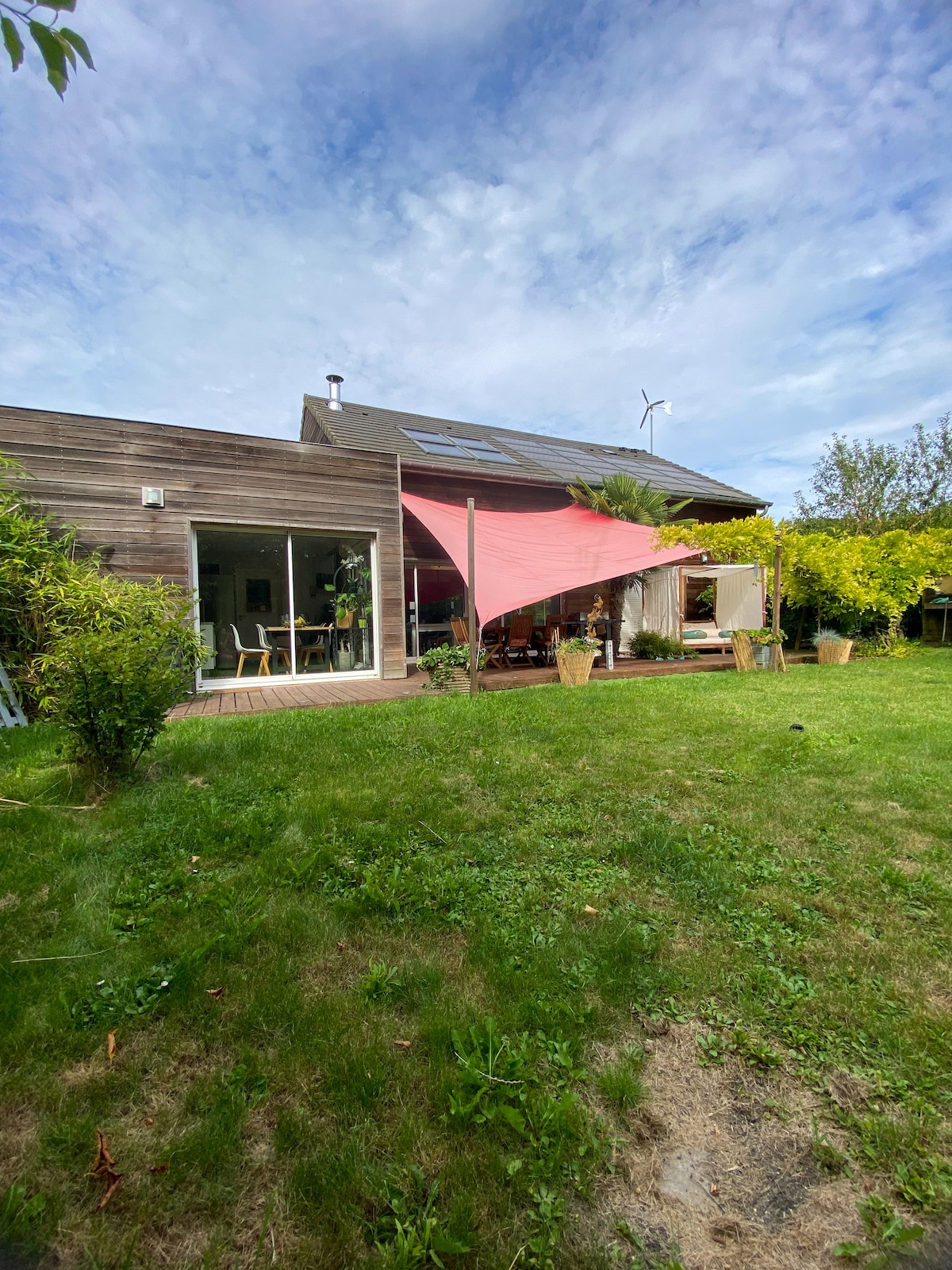 A spacious outdoor area is captured, featuring a green lawn bordered by various plants. A shaded terrace can be seen, with a vibrant red sail providing cover over dining areas. Large glass doors reveal the interior of the home, blending indoor and outdoor spaces.
