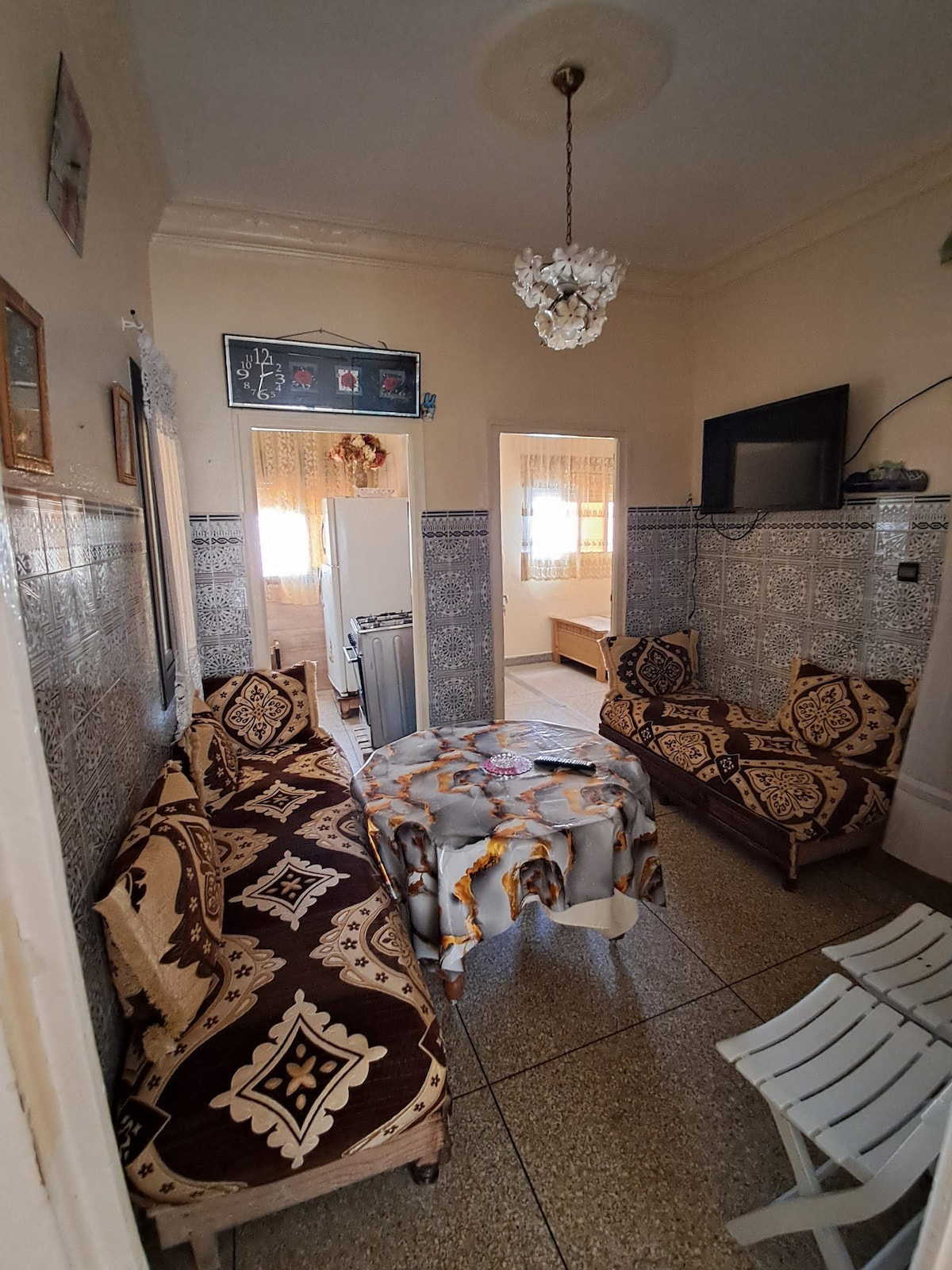 A cozy living area features patterned sofas surrounding a circular table. An old-fashioned chandelier hangs from the ceiling, and walls are adorned with decorative tiles. Light enters through several windows, with white curtains partially drawn, creating a welcoming ambiance in the space.