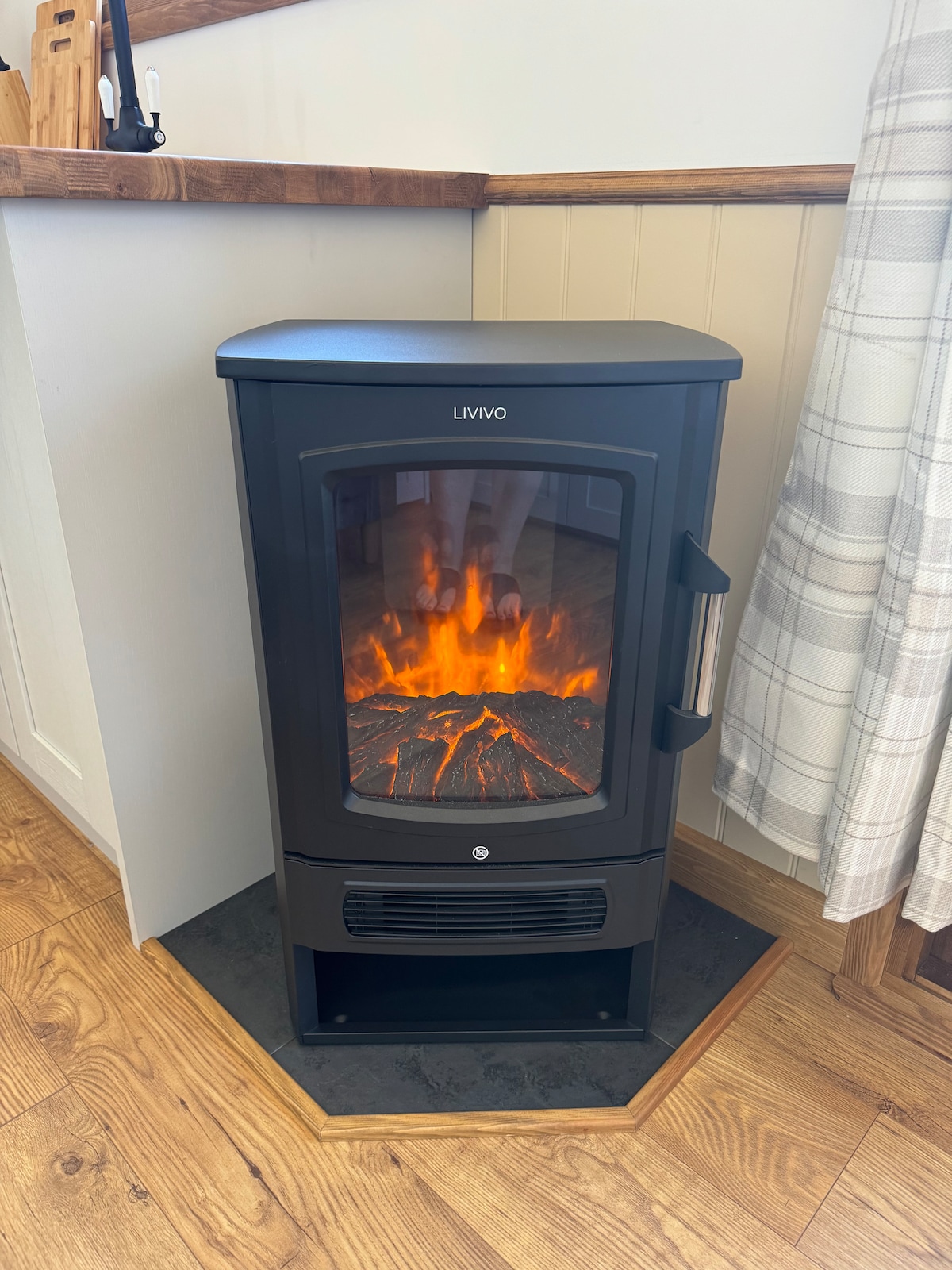 A modern black stove is positioned in the corner, featuring a realistic flame effect that adds warmth to the space. The surrounding area showcases light wooden flooring and a neutral wall, complemented by a soft-checkered blanket draped nearby.