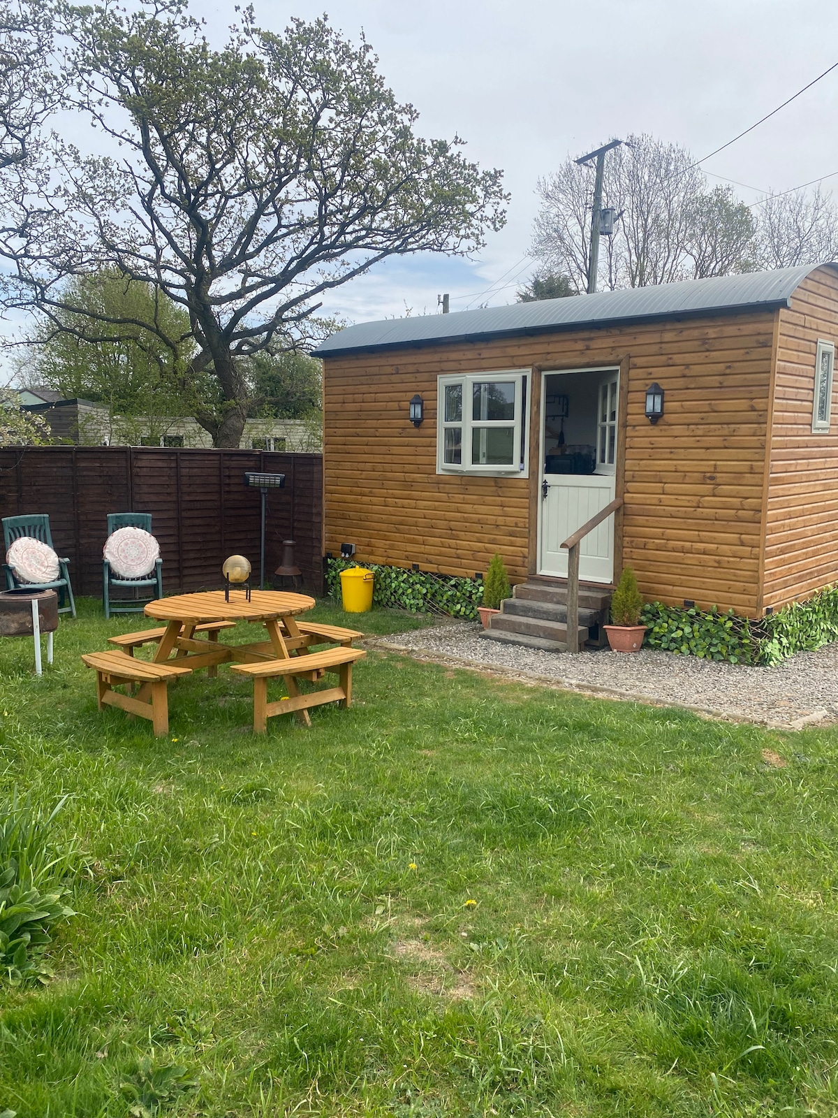 A charming exterior view of a wooden cabin is shown, featuring a small porch entry and surrounded by a grassy lawn. Two chairs and a picnic table are arranged in the outdoor area, with trees providing a natural backdrop.