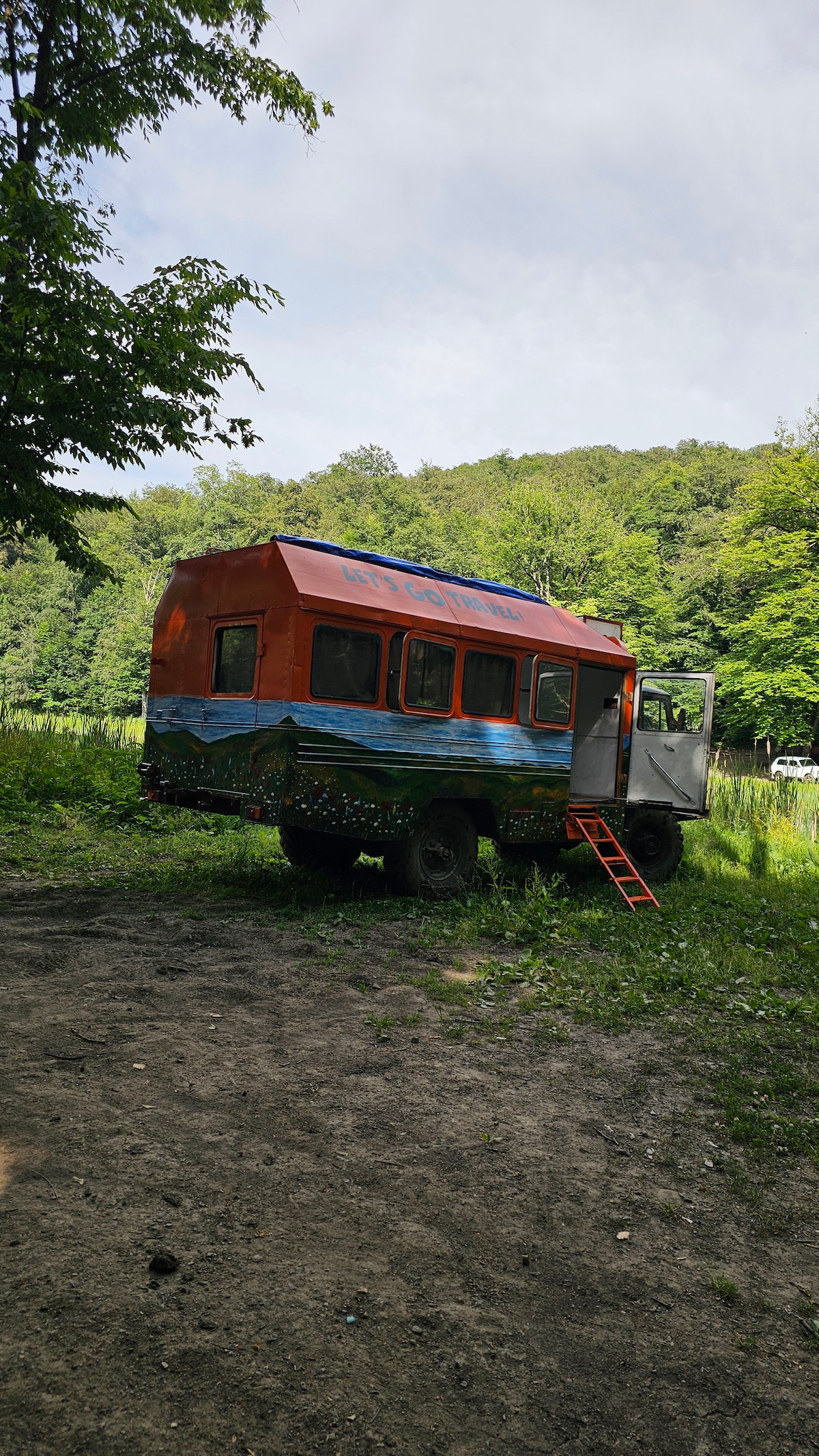 A vibrant orange camper van, decorated with nature-themed artwork, stands on a gravel patch surrounded by lush greenery. A small ladder leads to the entrance, and soft, natural light filters through the trees, creating a peaceful outdoor setting.