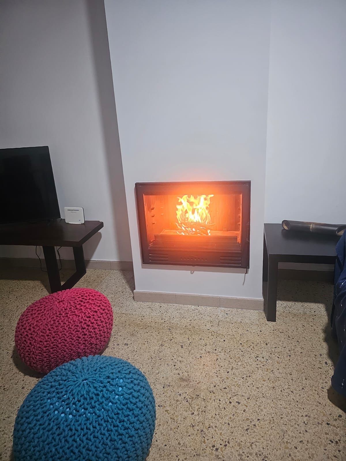 A modern fireplace is positioned against a smooth wall, with flames visible, providing a warm glow. Colorful poufs in pink and blue are placed nearby, alongside a dark wooden side table and a TV. The flooring features a light-colored texture.