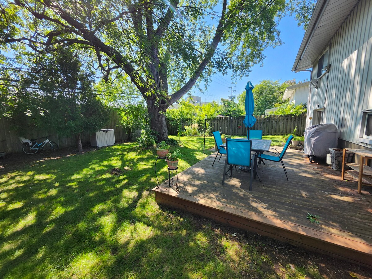 A spacious deck is situated under the shade of a large tree, surrounded by a well-kept lawn. Outdoor furniture with blue cushions is arranged around a table, complemented by a large umbrella. A grill is visible nearby, creating a comfortable space for outdoor gatherings.