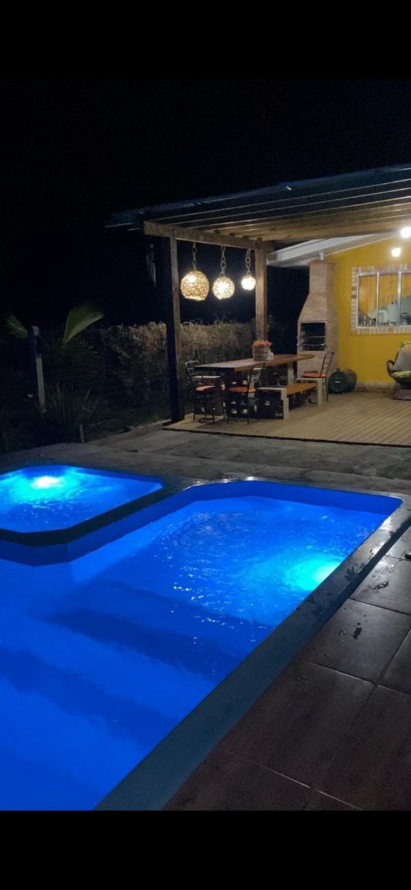 A well-lit outdoor area showcases a relaxing spa pool and a shallow pool, both illuminated with a calming blue light. Nearby, a dining table is set under a covered porch, accompanied by hanging lanterns that add a warm glow to the space.