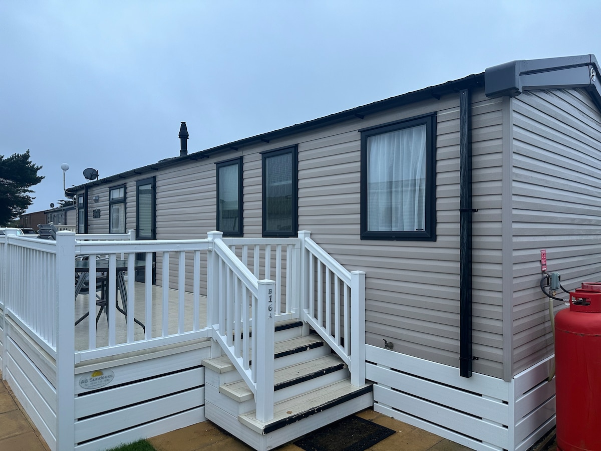A spacious caravan exterior is shown with a light-colored facade and several windows. A white staircase leads to a decked area, where outdoor furniture is visible. A red gas cylinder adds a pop of color, complementing the welcoming entrance.