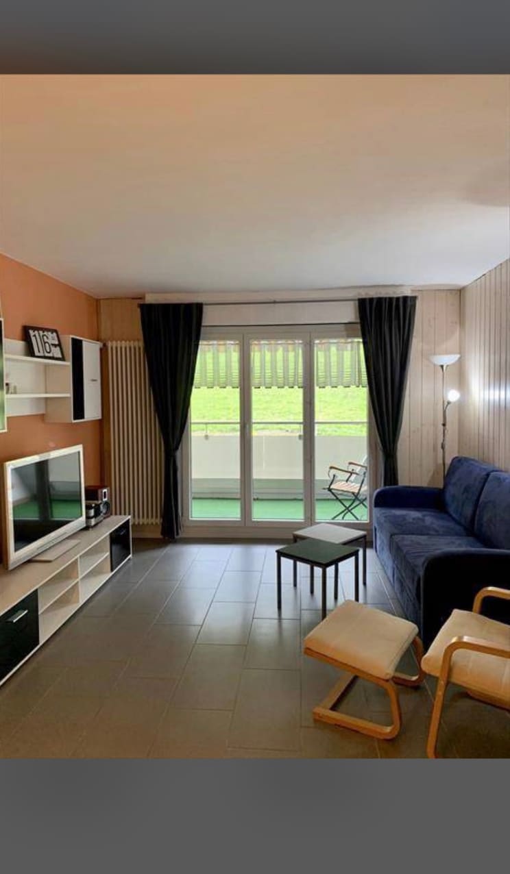 A living area features a blue sofa, a TV unit with a flat-screen television, and modern decor. Large glass doors lead to a balcony, complemented by light-filtering curtains. A small chair and a wooden stool are positioned nearby, providing additional seating options.