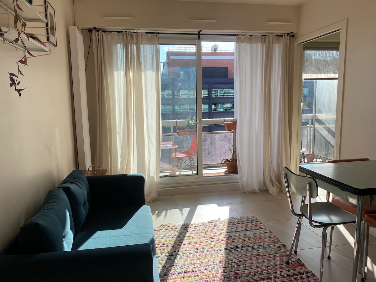 A modern living area features a spacious blue sofa positioned adjacent to large windows. Light curtains frame the windows, allowing natural light to illuminate the room. A colorful area rug lies on the tiled floor, and a dining table with chairs is nearby.