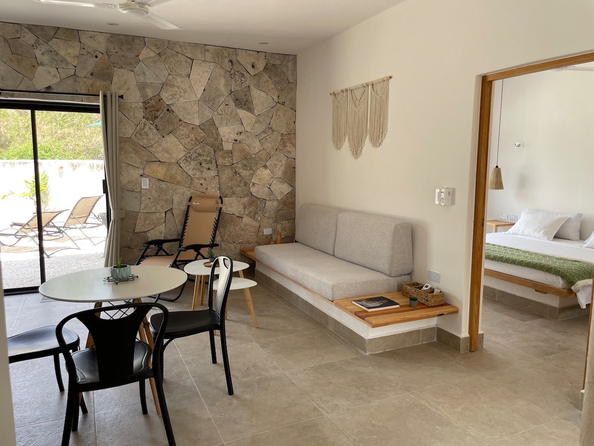 An open living space features a light-colored sofa against a textured stone wall. A round dining table with black chairs is visible. Sliding glass doors lead to an outdoor area with sunlight. A bedroom area is seen in the background, with a bed dressed in neutral tones.