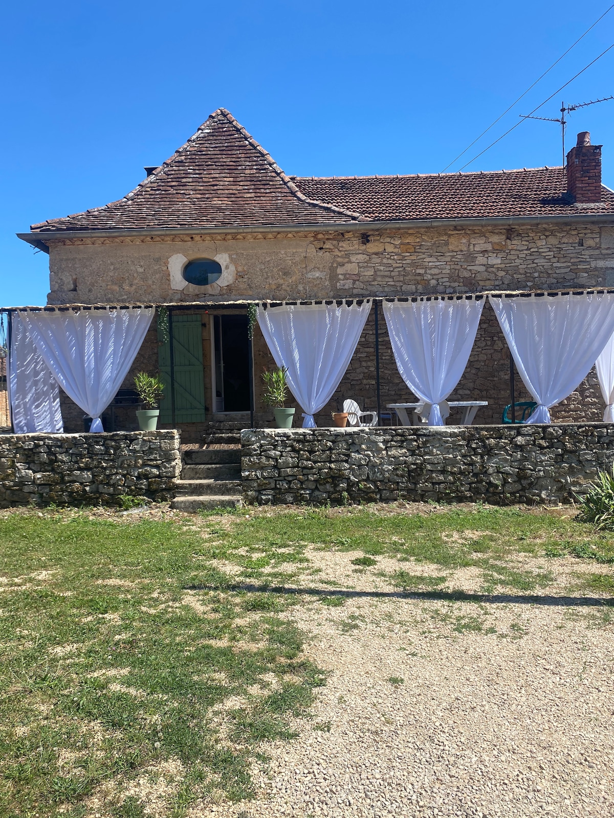 A rustic stone building features elegant white curtains draped along the terrace, framing the entrance. Potted plants add greenery to the space, while a set of outdoor seating is visible on the gravel path. The clear blue sky provides a bright backdrop.