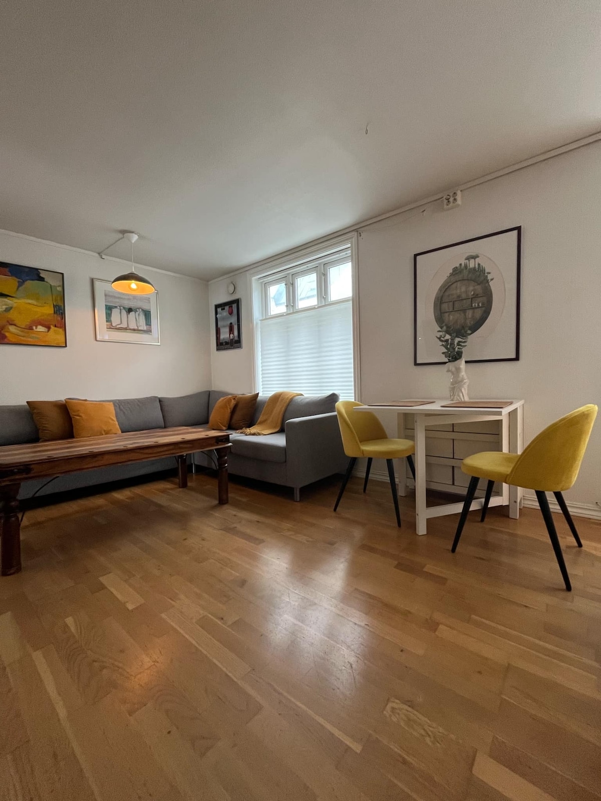A light-filled living area features a wooden table surrounded by yellow chairs and a gray sofa adorned with colorful cushions. Artwork is displayed on the walls, and sunlight streams through large windows, complementing the warm wooden floors.