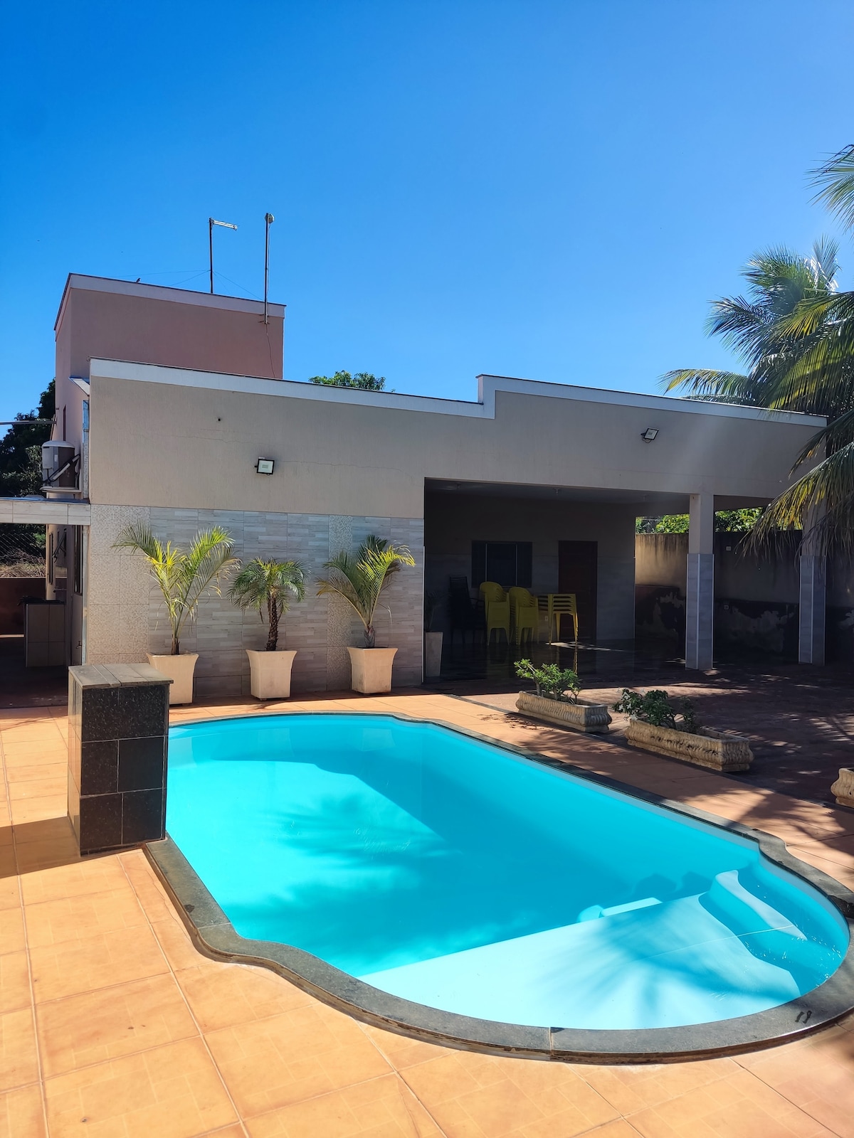 A well-maintained outdoor area features a clear blue pool surrounded by potted plants. The adjacent building showcases a covered patio area with yellow chairs visible, complemented by sunny skies above.