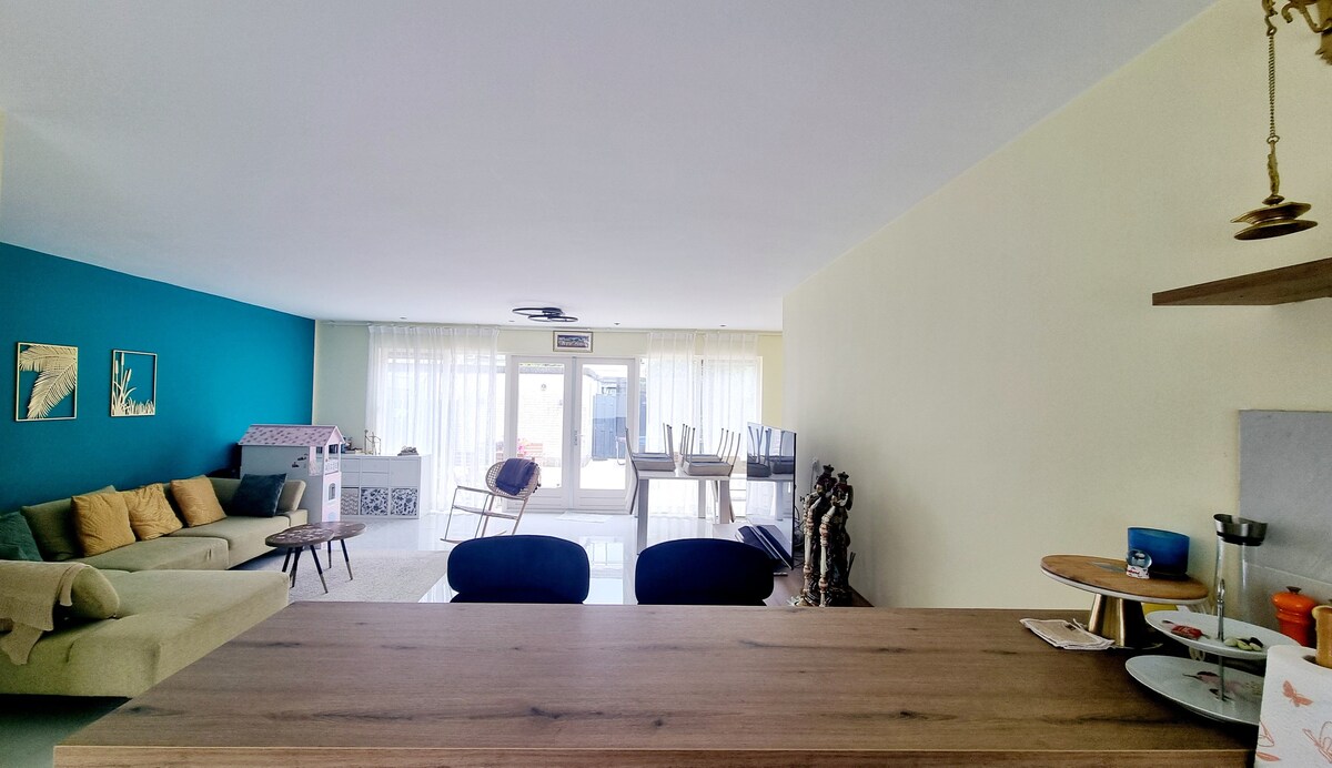A spacious living area showcases a mix of neutral and vibrant colors, with a large window allowing natural light to fill the room. A wooden countertop separates the living space from the kitchen, while a cozy sofa and decorative elements invite relaxation.