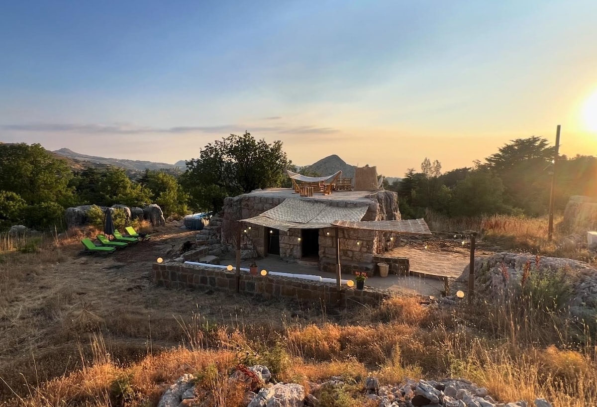 A unique guesthouse is nestled among the cedar trees, featuring a natural stone exterior. An outdoor seating area with wooden furniture is visible, surrounded by lush greenery. Relaxing loungers are positioned nearby, with a scenic landscape and sunset view in the background.
