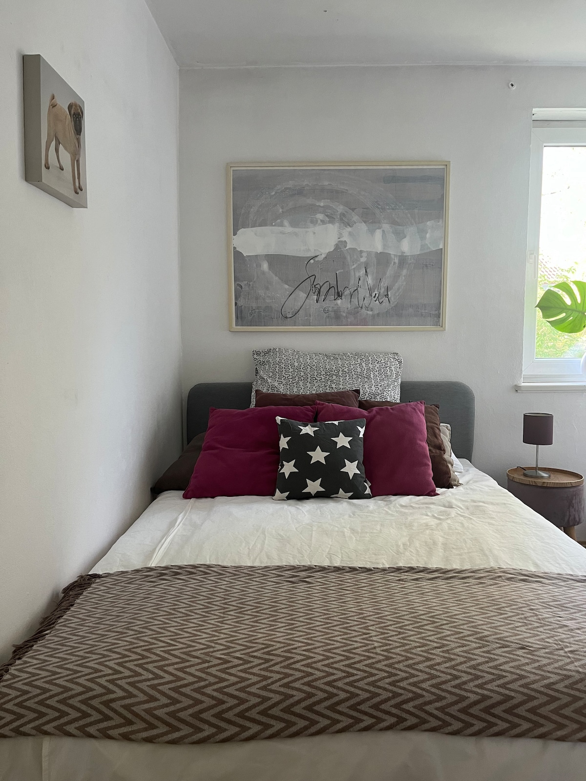 A cozy sleeping area features a single bed dressed with layered bedding in neutral tones and patterned cushions. A piece of artwork hangs above the bed, and a small lamp sits on a bedside table. Natural light enters through a nearby window, illuminating the space.