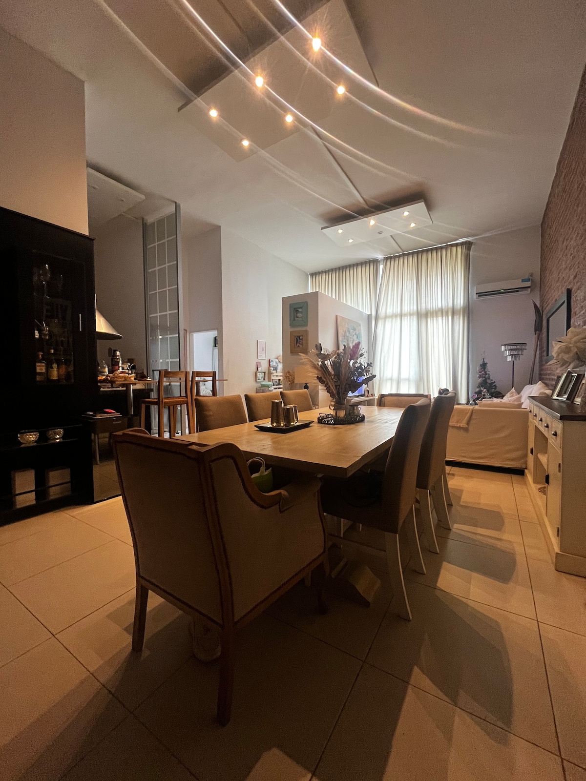A spacious dining area features a wooden table surrounded by beige and white chairs. Soft lighting from hanging fixtures complements the large windows dressed with sheer curtains, allowing natural light to brighten the room. A subtle decorative arrangement sits on the table.