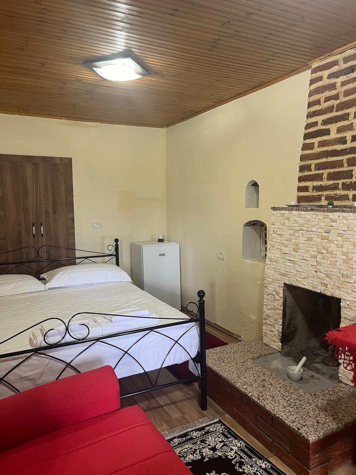 A double bedroom is featured, showcasing a spacious bed with white linens. A small fridge is located against one wall, while a fireplace adds warmth to the room. A red sofa is positioned near the bed, contributing to the overall comfort of the space.