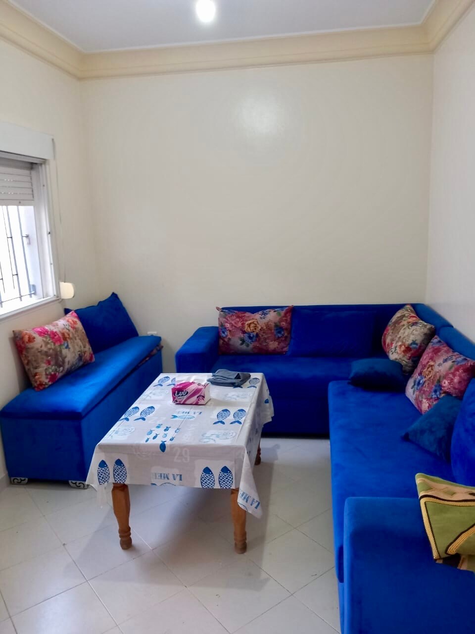 A cozy living area is furnished with a bright blue sectional sofa, complemented by colorful decorative pillows. A simple table covers a patterned tablecloth, and natural light filters through a window, creating a pleasant ambiance in the space.