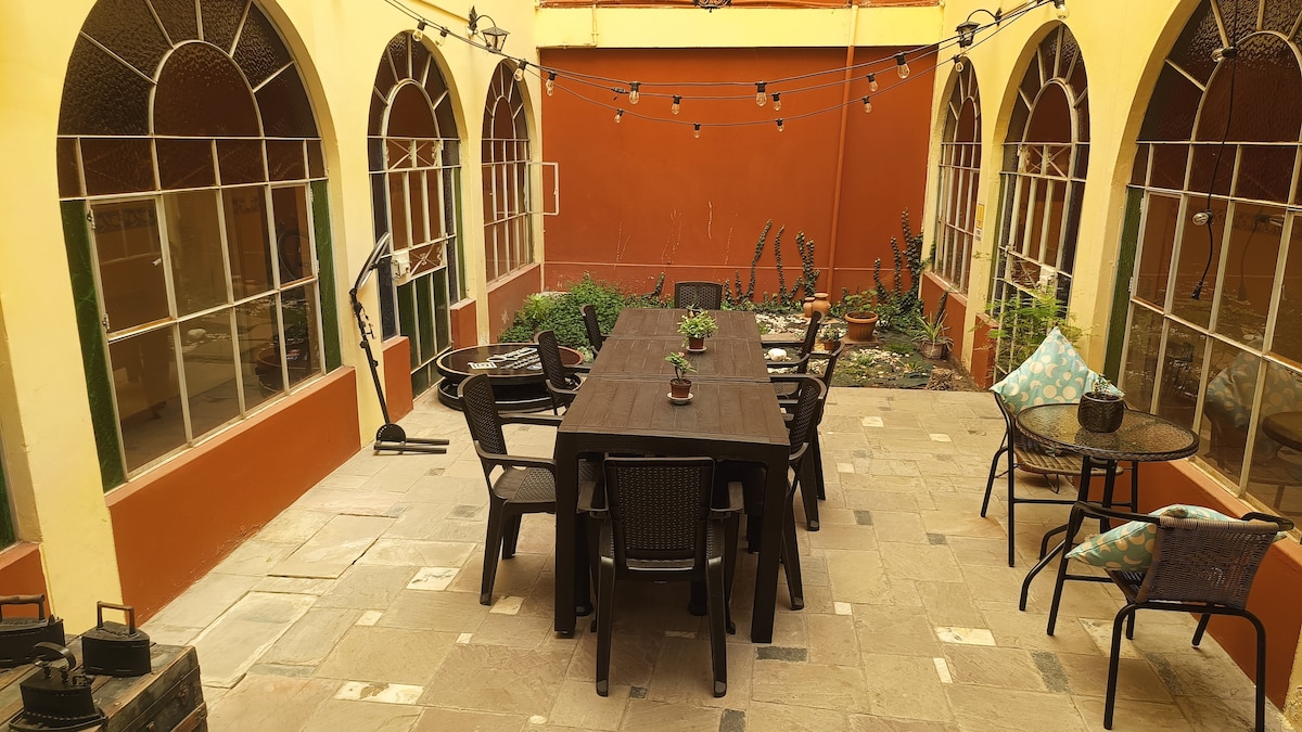 An outdoor courtyard is featured, showcasing a long, dark wooden table surrounded by multiple chairs. Potted plants are placed throughout the space, with string lights overhead. The walls are painted in warm tones, complemented by large arched windows framing the area.