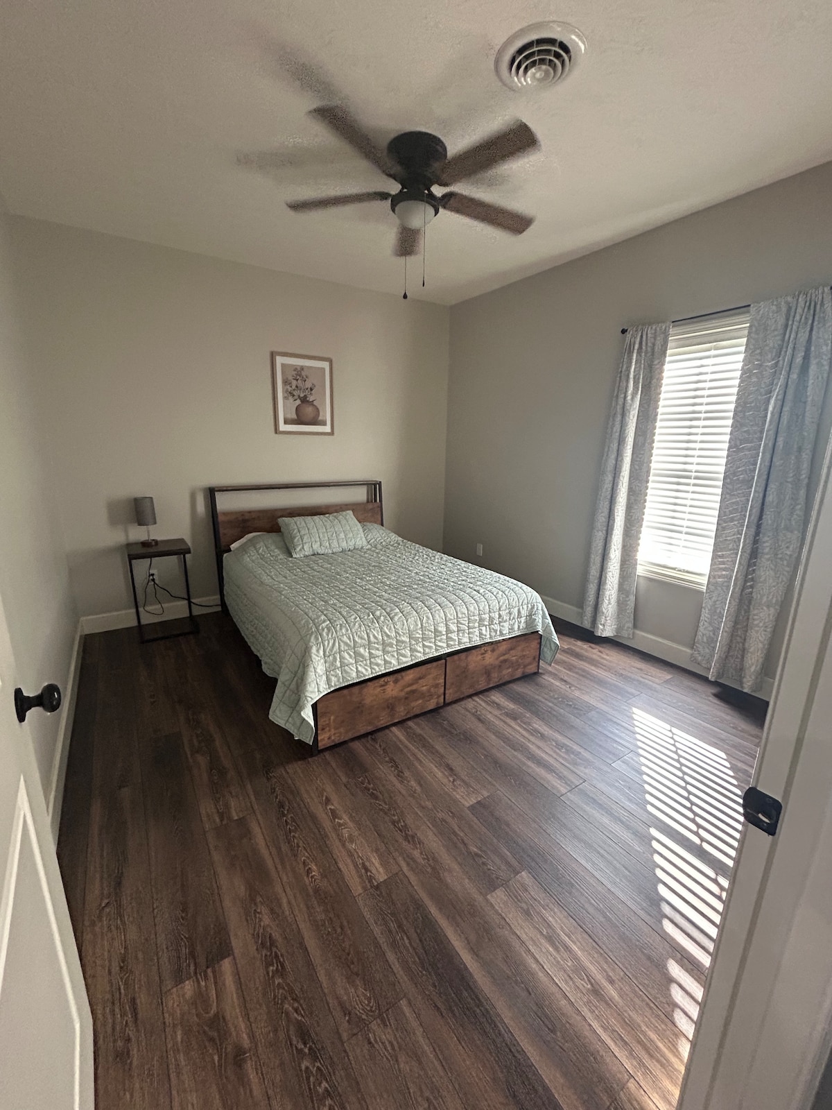 A subtly decorated bedroom features a wooden bed with a light quilt and a pillow. A nightstand holds a lamp beside the bed. Natural light filters through a window with light curtains, casting patterns on the wooden floor. A ceiling fan completes the space.