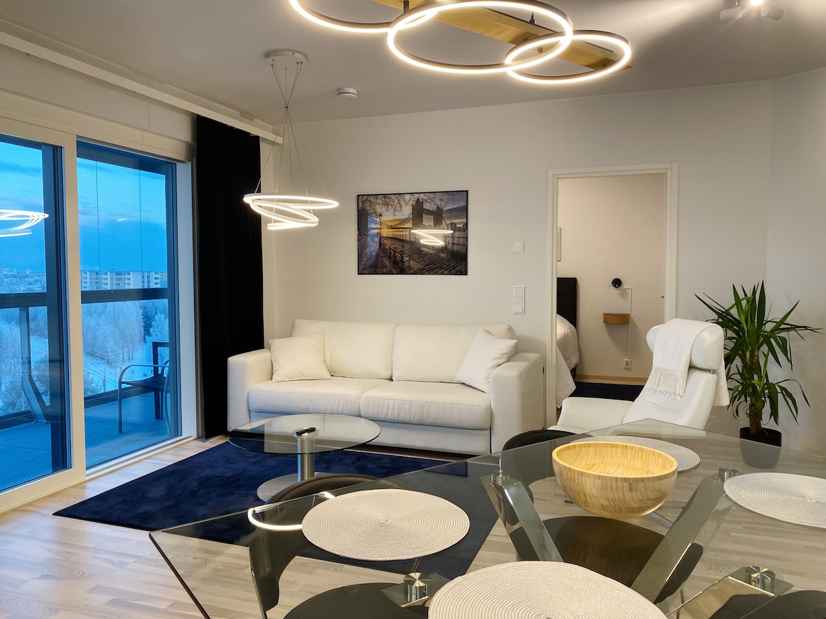 The living area includes a white sofa and a glass dining table set with round placemats. A stylish light fixture hangs above, and an indoor plant is positioned near a door leading to another room. Large windows provide views of the city and the sea.