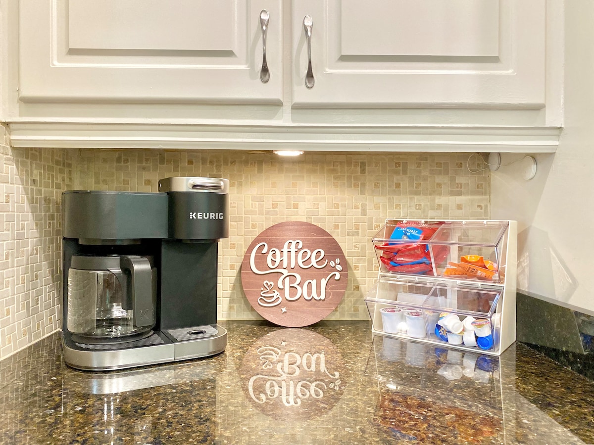 A coffee bar area is visible, featuring a Keurig coffee maker and a glass-front organizer filled with assorted coffee pods and creamers. A decorative sign stating 'Coffee Bar' adds a charming touch to the counter, with neutral-colored tiles providing a backdrop.