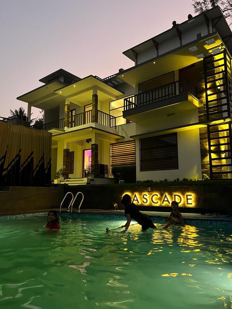 A sleek two-story house is illuminated by soft golden lights in the evening. The inviting pool reflects the warm glow, with several figures enjoying the water. A sign reading 'Cascade' adds a modern touch to the outdoor area surrounded by lush greenery.
