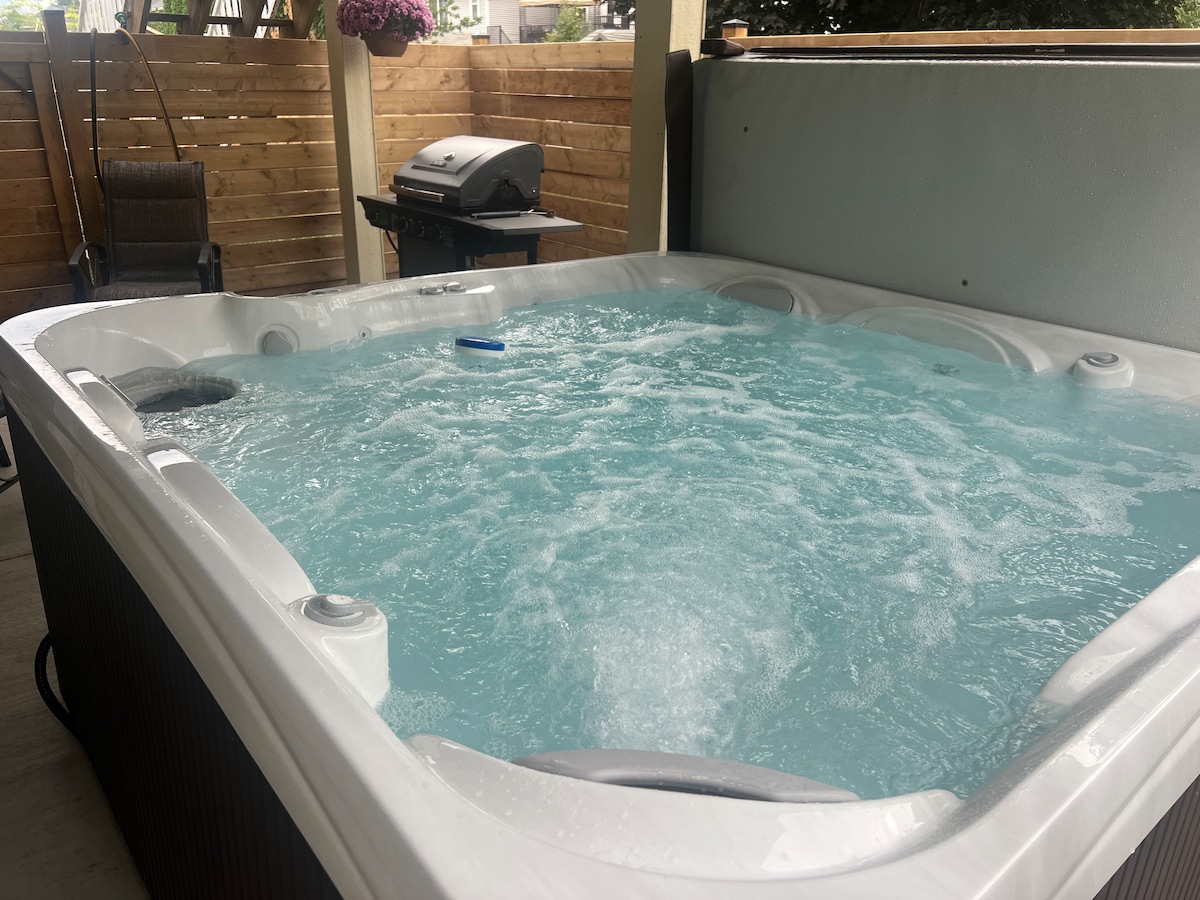 A private hot tub is prominently featured on the outdoor patio. The bubbling water reflects a soothing blue hue, surrounded by a fenced garden area. A propane barbeque is positioned nearby, creating a relaxing atmosphere for unwinding after a day of activities.