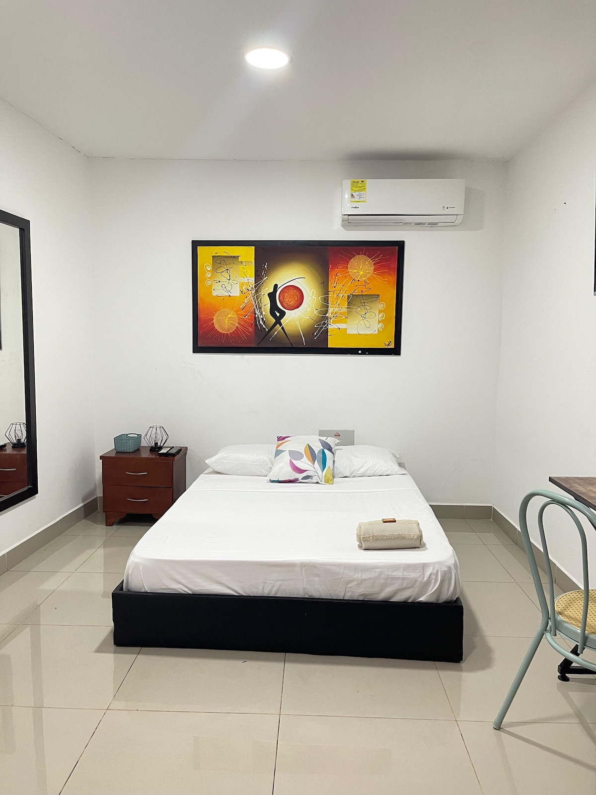 A simple sleeping area features a spacious bed with fresh linens and a towel neatly folded. A colorful wall art piece adds interest above the bed. A nightstand is positioned beside the bed, while a small table and chair occupy one corner of the room. Natural light is welcomed through a window.