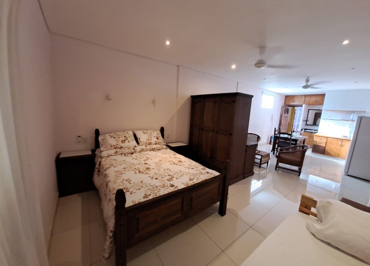 A spacious studio is shown, featuring a wooden bed with floral bedding, accompanied by matching nightstands. A large cabinet is positioned beside the bed. Light-coloured tiles reflect the illumination from overhead lights, while a kitchenette area with dining set is visible in the background.