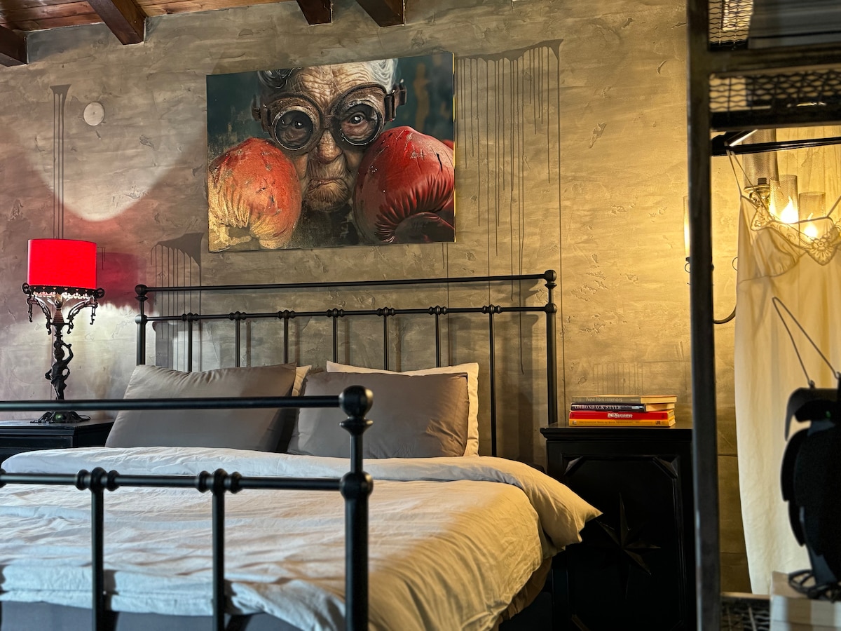 A stylish bedroom features a spacious bed with a light-colored comforter and matching pillows. A piece of wall art is displayed above the bed, while a red lamp casts a warm glow. The room is adorned with a textured gray wall and exposed wooden beams.