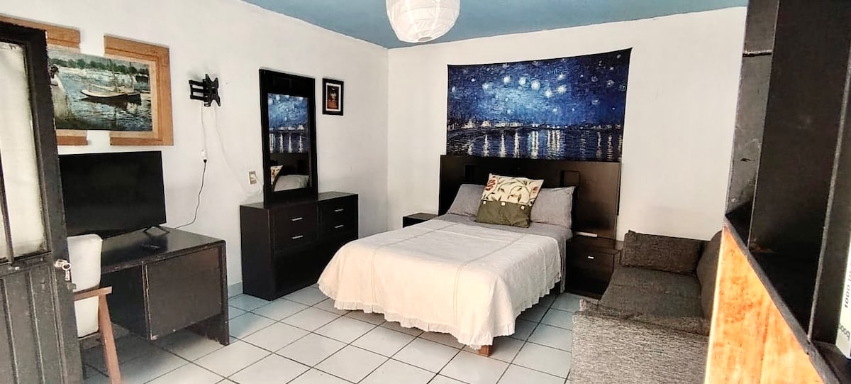 A bright and airy room is furnished with a spacious bed dressed in neutral linens. A wall-mounted HD TV is positioned across from the bed. A large piece of artwork depicting a night sky adds interest, while dark wood furniture provides storage and functionality.